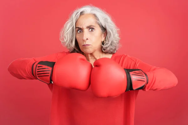 Studio portrait with red background of a rude mature woman in boxing gloves looking at camera