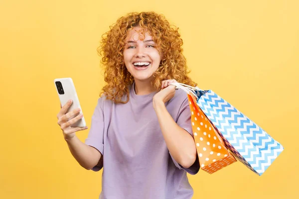 stock image Studio image with yellow background of a young smiley woman with curly hair using the mobile while holing shopping bags