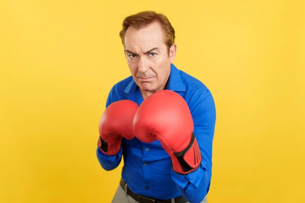 Mature man with red boxing gloves in guard pose of boxing in studio with yellow background