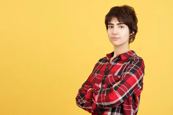 Serious androgynous person looking at camera with arms crossed in studio with yellow background