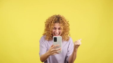 Studio video with yellow background of an excited woman with curly hair celebrating while using the mobile phone