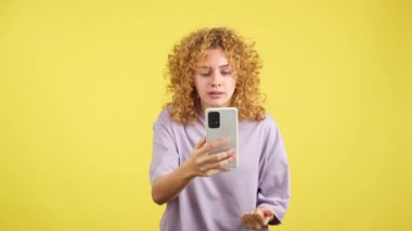 Studio video with yellow background of a upset and worried woman with curly hair using a mobile
