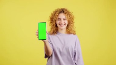 Studio video with yellow background of a smiley woman looking at camera while holding a mobile with the chroma screen