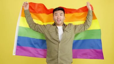 Happy chinese man raising a lgbt rainbow flag in studio with yellow background