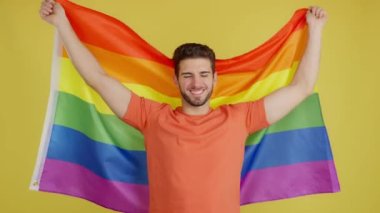 Happy caucasian man raising a lgbt rainbow flag in studio with yellow background