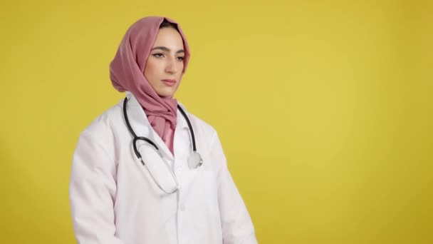 深刻な女性イスラム教徒医師とともに腕交差見ます離れてスタジオでともに黄色の背景 — ストック動画