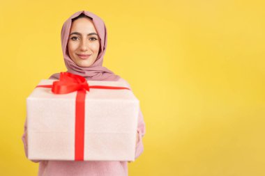 Stüdyoda sarı arka planda bir hediye verirken kameraya gülümseyen mutlu Müslüman kadın.