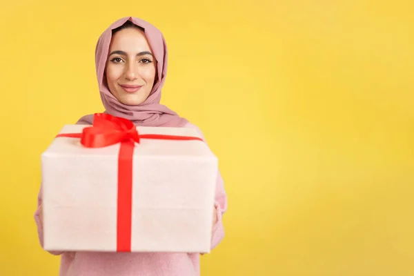 幸せなイスラム教徒の女性は黄色の背景を持つスタジオでの贈り物を与えながら カメラで笑顔 — ストック写真