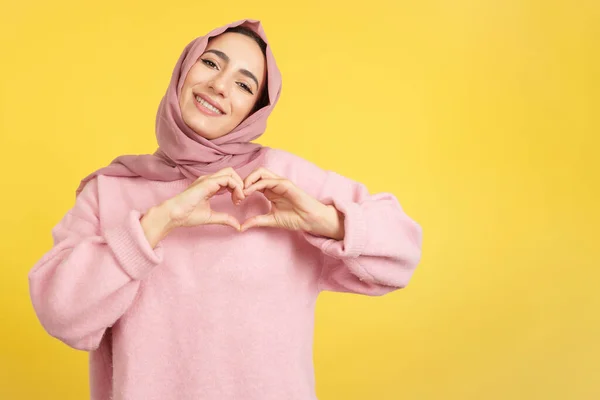 黄色の背景を持つスタジオで指の心の形を表すイスラム教徒の女性 — ストック写真
