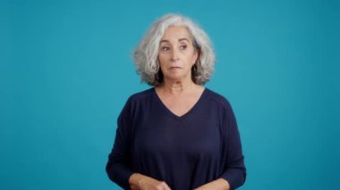 Elleri panik içinde hareket eden olgun bir kadının mavi arka planlı stüdyo videosu.