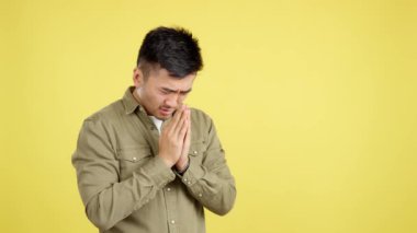 Çinli dua eden bir adam, elleri bağlı, sarı arka planda stüdyoya bakıyor.