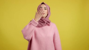 Stüdyoda sarı arka planda kameraya bakan Müslüman bir kadın öpüşüyor.