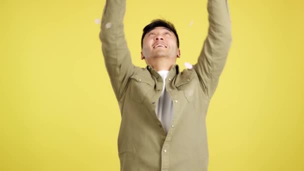 快乐的中国男人被五彩纸屑环绕着 在黄色背景的演播室里在空中飞舞 — 图库视频影像