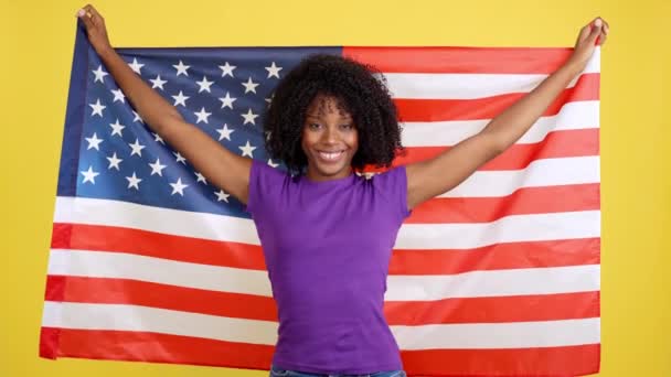 黄色の背景を持つスタジオで米国のフラグを上げるアフロ髪を持つ幸せな女性 — ストック動画
