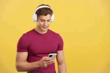Stüdyoda telefon ve kulaklıklarla müzik dinleyen, sarı arka planlı, rahat, beyaz bir adam.