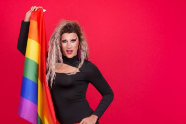 Travesti kılığındaki travesti stüdyoda kırmızı arka planı olan gökkuşağı lgbt bayrağıyla kameraya bakıyor.