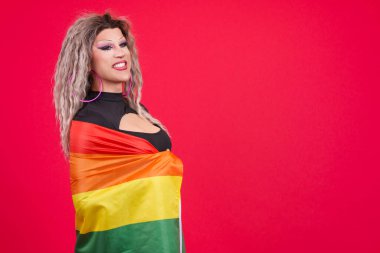 Travestiler gibi giyinmiş gururlu bir transseksüel stüdyoda kırmızı arka planlı gökkuşağı lgbt bayrağıyla sarılıyor.