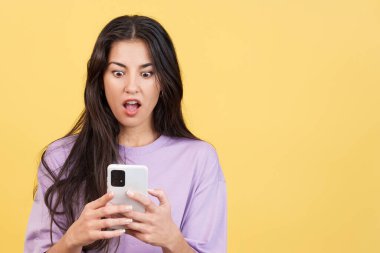 Stüdyoda sarı arka planda bir cep telefonunun ekranına bakan sürpriz bir kadın.