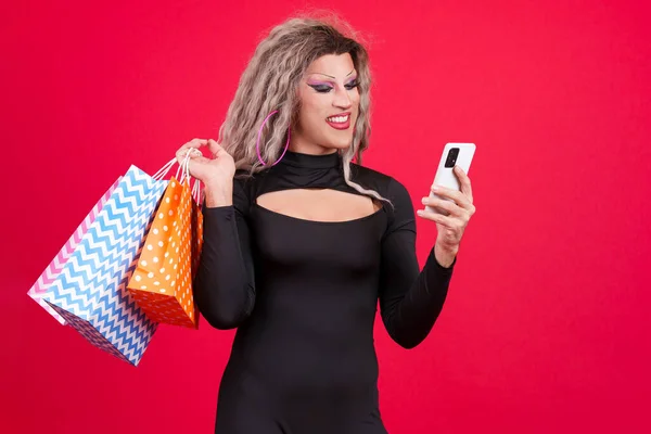 transgender person dressed as drag queen using a mobile while holding shopping bags in studio with red background
