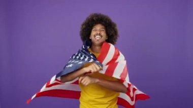 Kuzey Amerika ulusal bayrağıyla sarılı kıvırcık saçlı mutlu Afrikalı adam stüdyoda kameraya bakıyor.