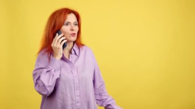 Kızgın kızıl saçlı olgun kadın sarı arka planlı cep telefonuyla konuşuyor.