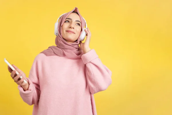 黄色の背景を持つスタジオで携帯電話やヘッドフォンで音楽を聴きながら笑顔幸せなイスラム教徒の女性 — ストック写真