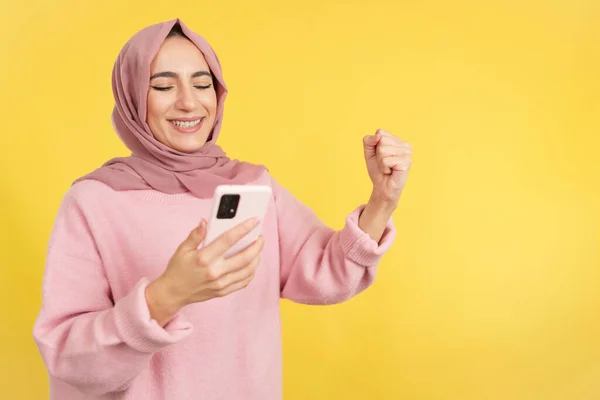 黄色の背景を持つスタジオでモバイルを使用している間に拳を上げ興奮したイスラム教徒の女性 — ストック写真