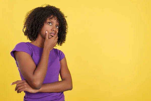 Woman with afro hair standing with thoughtful expression in studio with yellow background