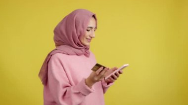 Stüdyoda sarı arka planda alışveriş yapmak için cep telefonu ve kartı kullanan Müslüman bir kadın.