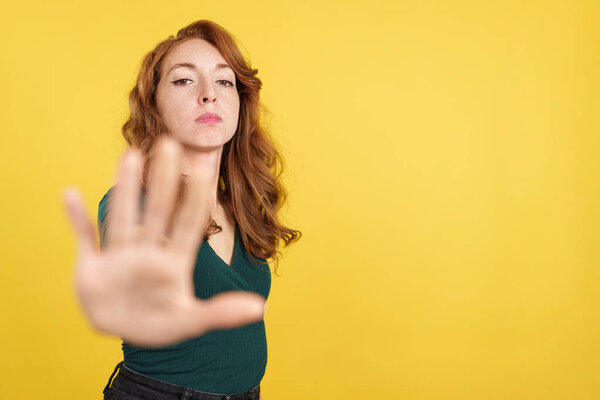 Redheaded woman gesturing prohibition with hand in studio with yellow background