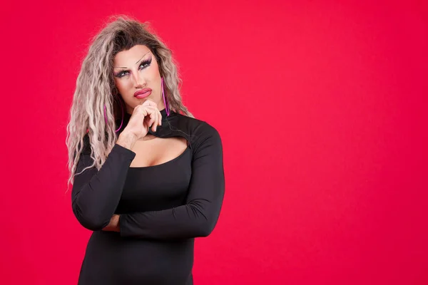 Transgender person with black dress and make up with hand on chin and thoughtful expression in studio with a red background
