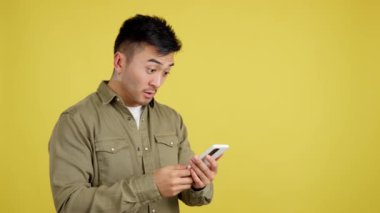 Surprised chinese man looking the screen of a mobile in studio with yellow background
