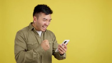 Stüdyoda sarı arka planda cep telefonunu kullanarak kutlama yapan heyecanlı Çinli adam.