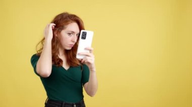 Saçına dokunan kızıl saçlı kadın cep telefonuna yansıyor. Sarı arkaplan