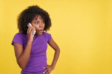 Afro saçlı bir kadın stüdyoda sarı arka planlı kızgın bir ifadeyle cep telefonuyla konuşuyor.