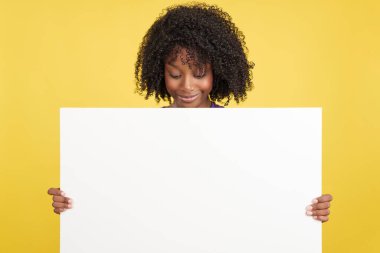Afro saçlı bir kadın sarı arka planlı stüdyosunda elinde tuttuğu boş bir tahtaya bakıyor.