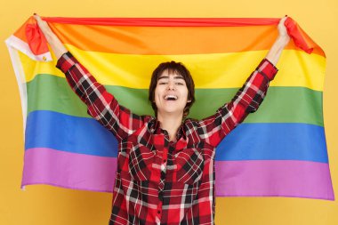 Androgynous sarı arka planlı stüdyoda bir lgbt gökkuşağı bayrağı göndere çekiyor.