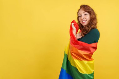 Stüdyoya lgbt bayrağı sarılmış, sarı arka planı olan mutlu kızıl saçlı kadın.