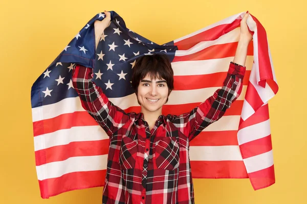 An androgynous person raising a USA flag in studio with yellow background