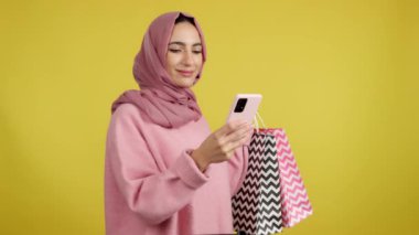 Müslüman bir kadın alışveriş torbalarını deliğe sokarken cep telefonunu kullanıyor. Sarı arkaplan