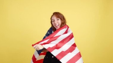 Stüdyoda sarı arka planda Kuzey Amerika bayrağıyla sarılan mutlu kızıl saçlı kadın.