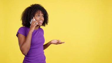 Angry woman with afro hair standing while talking to the mobile phone in studio with yellow background
