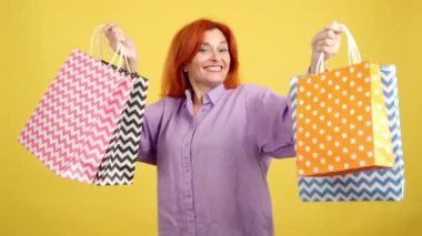 Stüdyoda sarı arka planda kameraya gülümserken bir sürü alışveriş torbası kaldıran olgun kızıl kadın.