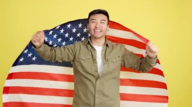 Mutlu Çinli adam kameraya bakıyor ve sarı arka planda Kuzey Amerika ulusal bayrağıyla sarılıyor.