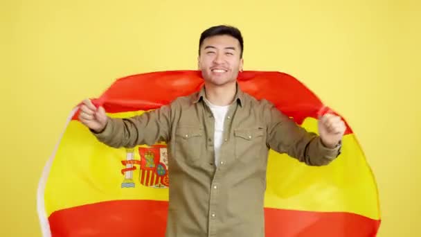 Szczęśliwy Chińczyk Patrzący Kamerę Owiniętą Hiszpańską Flagą Narodową Studio Żółtym — Wideo stockowe