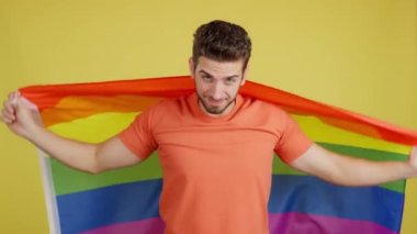 Stüdyoda sarı arka planda lgbt bayrağıyla sarılı mutlu beyaz adam.