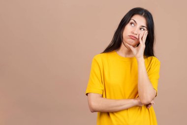 Sıkılmış beyaz kadın ayakta dikiliyor ve kahverengi arka planlı stüdyoya bakıyor.