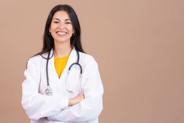 Kameraya gülümseyen beyaz kadın doktor. Stüdyoda kollarını kavuşturup kahverengi arka planda.