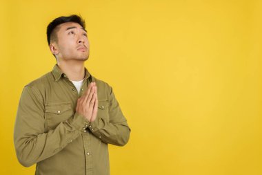 Çinli dua eden bir adam, elleri bağlı, sarı arka planda stüdyoya bakıyor.