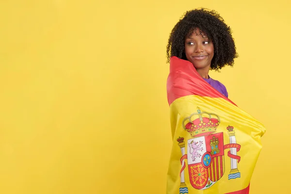 Distracted migrant afro woman wrapped with a Spanish flag while looking away in studio with yellow background